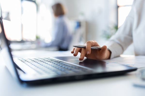 A person’s hand holding a pen hovers over a laptop touchpad on a bright desk, with another person blurred in the background.