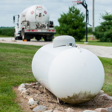 A large white propane tank sits on a gravel bed in a grassy yard, perfect for capturing in a short-form video, with a propane delivery truck parked on the driveway near some trees in the background.