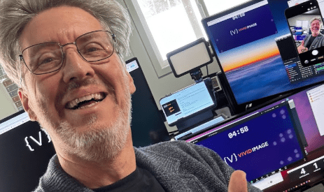 Smiling man with glasses gives a thumbs up in an office with several monitors, one of which displays his image. Daylight streams in from a window behind him.