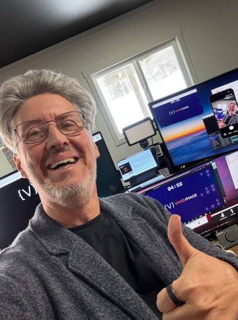 Smiling man with glasses gives a thumbs up in an office with several monitors, one of which displays his image. Daylight streams in from a window behind him.
