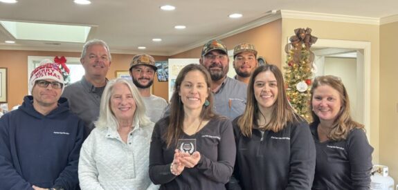 A group of nine people smiling and standing together indoors, some wearing festive hats. A Christmas tree decorated with lights and ornaments is in the background. One woman in front holds a small award.