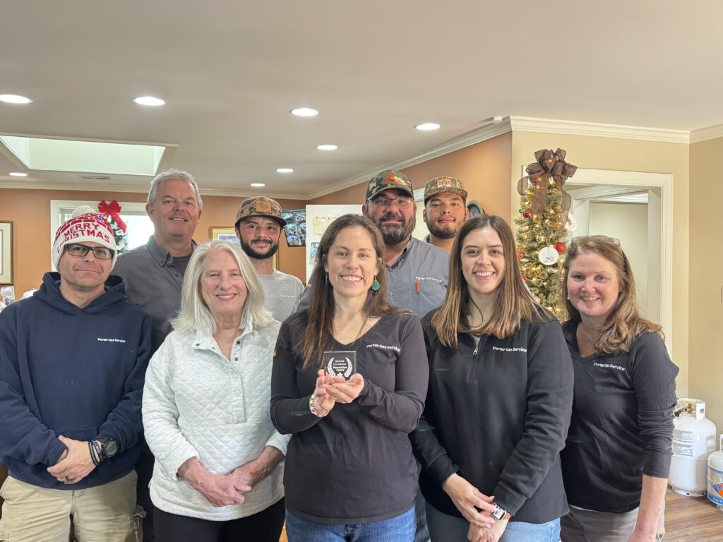 A group of nine people smiling and standing together indoors, some wearing festive hats. A Christmas tree decorated with lights and ornaments is in the background. One woman in front holds a small award.