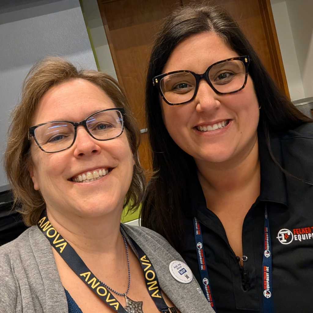 Two women in glasses and lanyards smile for an indoor selfie.