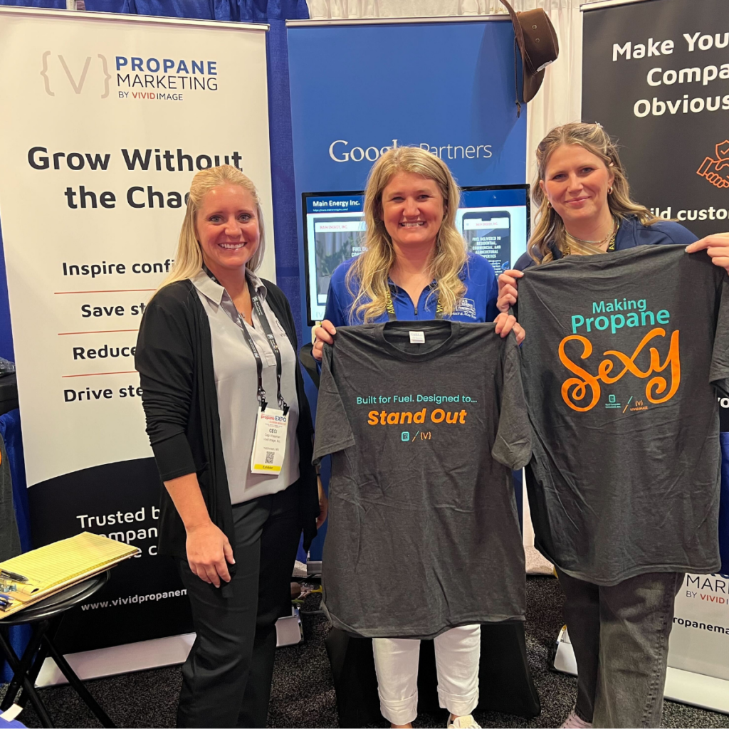 Three women at a propane booth; two hold T-shirts reading "Stand Out" and "Making Propane Sexy." Promo banners behind them.