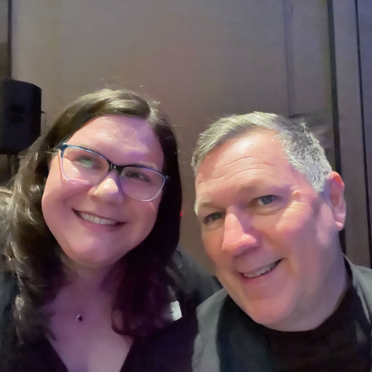 Two people indoors, one in glasses, smiling at the camera with a blurred background.