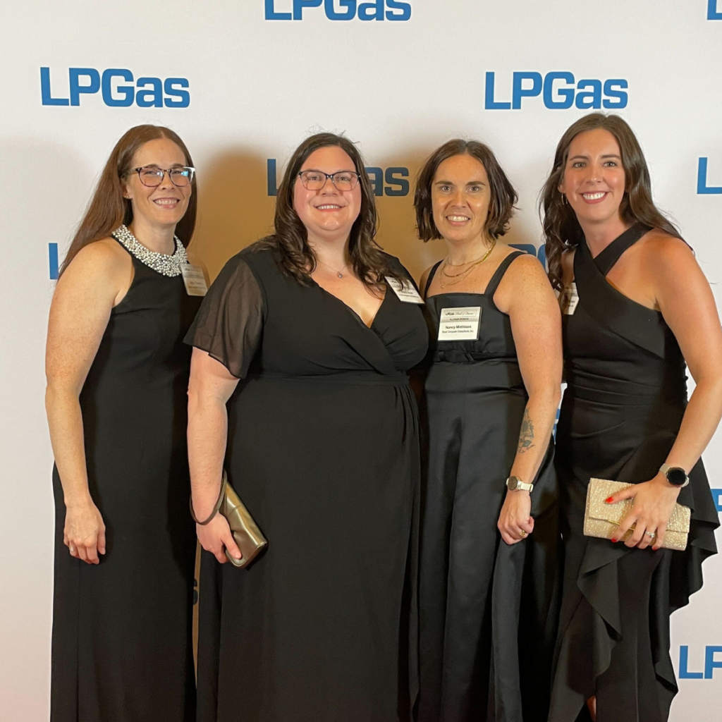 Four women in black dresses pose together before an LPGas logo backdrop at a formal event.