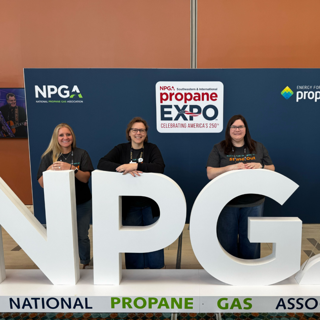 Three women stand behind large white NPG letters at the NPGA Propane Expo, with signage and logos in the background.
