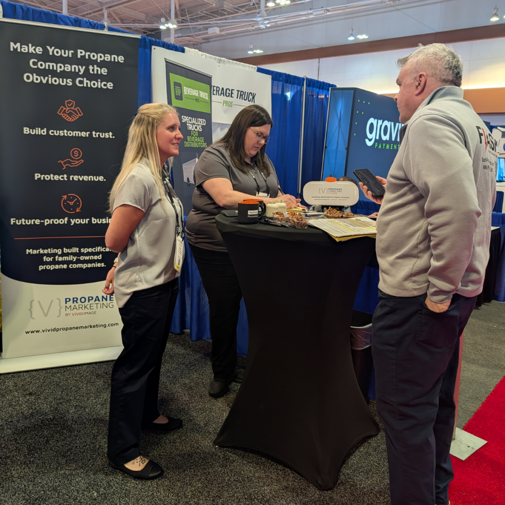 Two women at a marketing booth speak with a man at a trade show, display items and promos visible on the table.