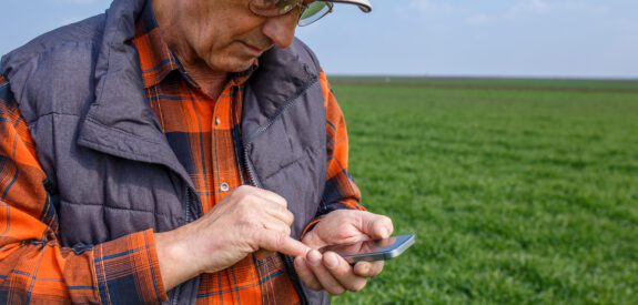 An older man in a cap and glasses stands in a green field, using his smartphone with one hand.