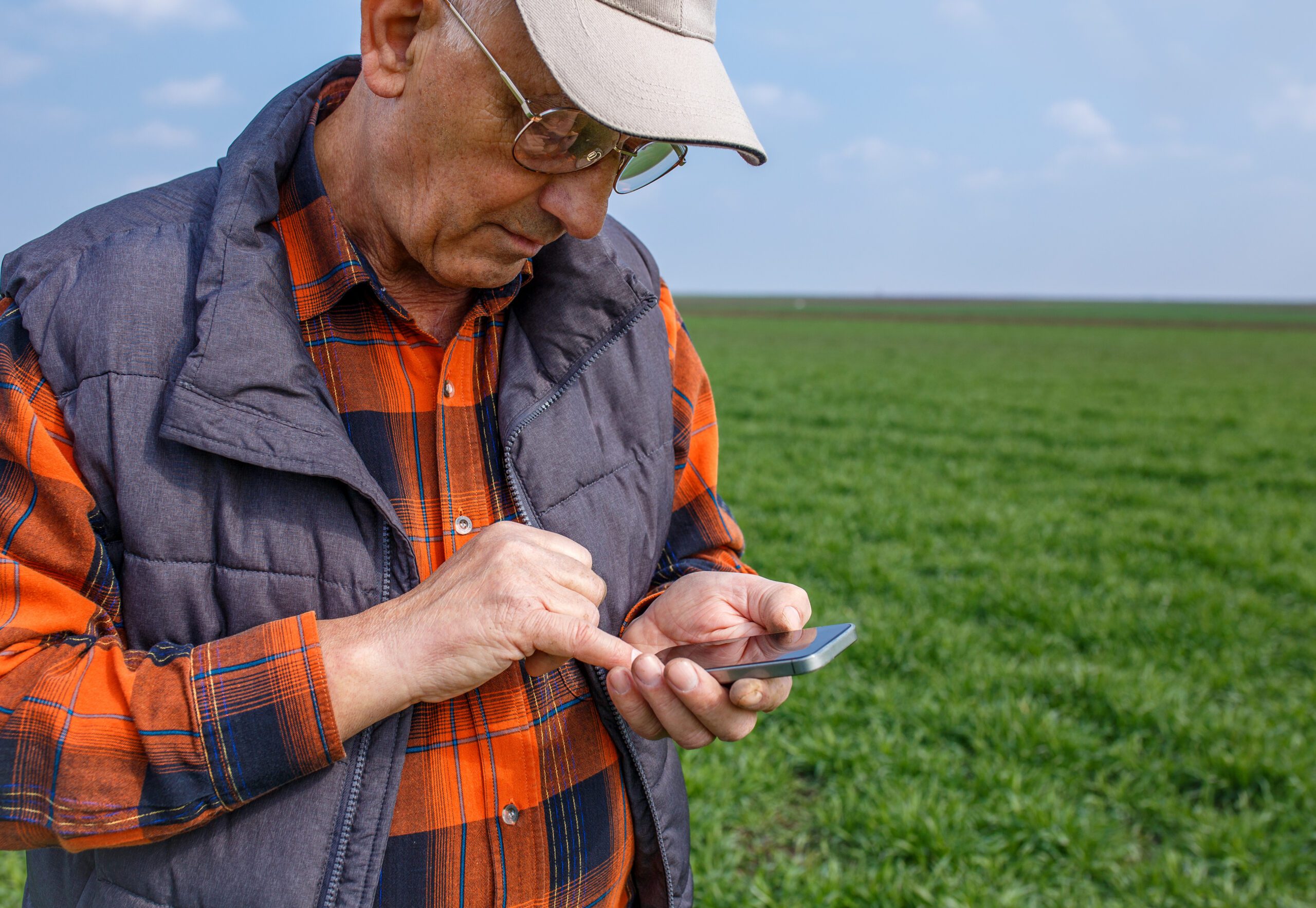 An older man in a cap and glasses stands in a green field, using his smartphone with one hand.
