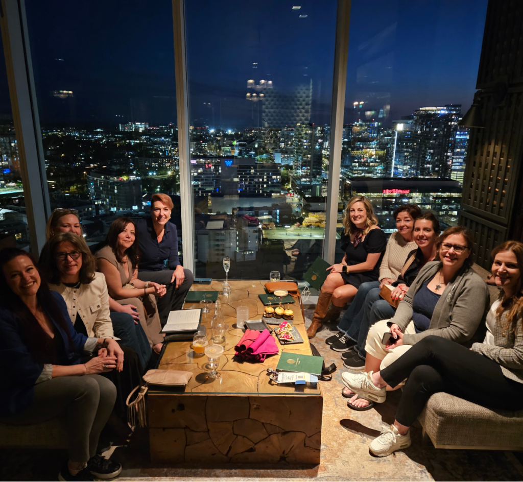 Eleven women gather around a table with drinks and snacks in a high-rise lounge, city lights glowing outside at night.