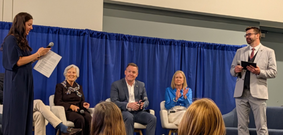 Five people onstage in a panel: two standing with mics, three seated, blue curtain backdrop, audience in front.