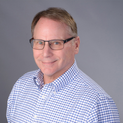 Smiling man in glasses and a blue-and-white checkered shirt stands before a plain gray background.