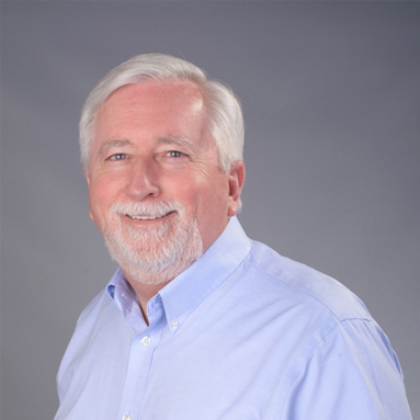 Smiling older man with white hair and beard in a light blue shirt, standing before a plain gray background.